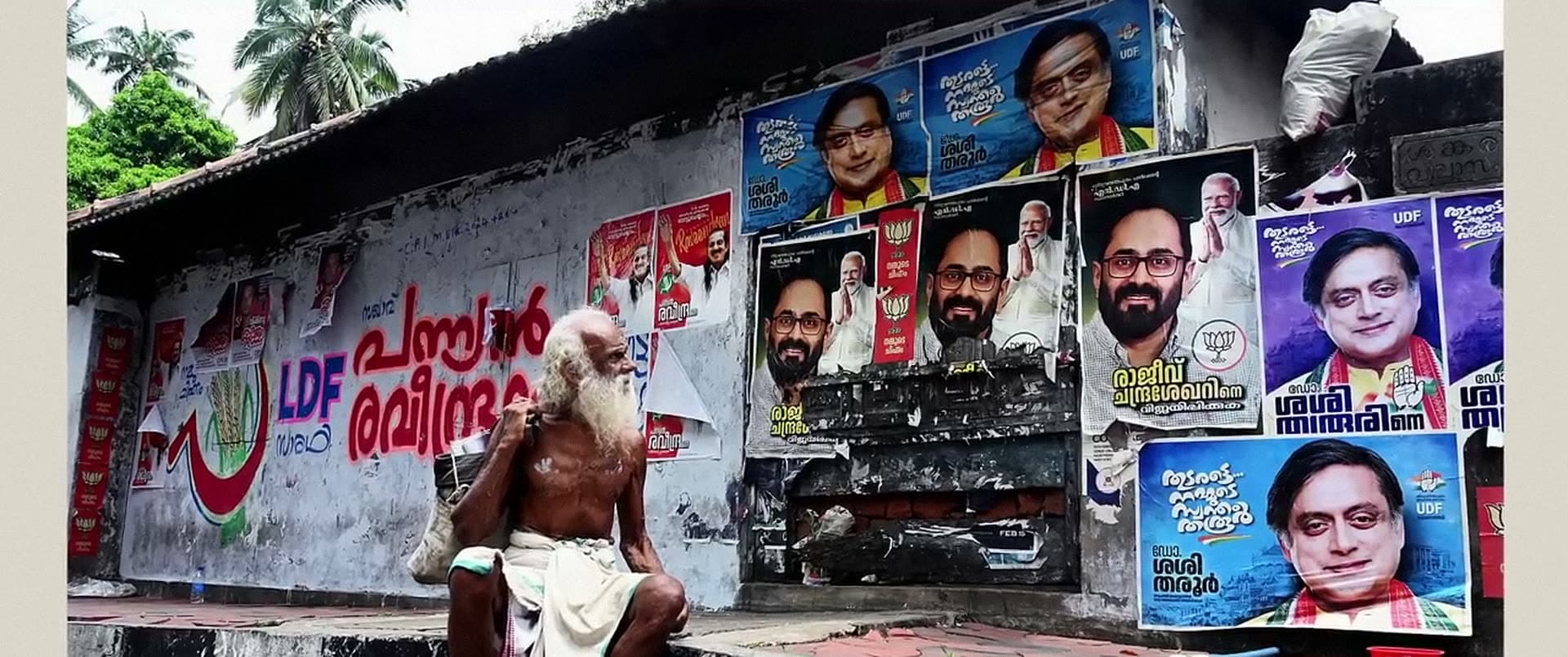 An elderly man sits on a ledge below a wall covered in painted slogans and election posters.