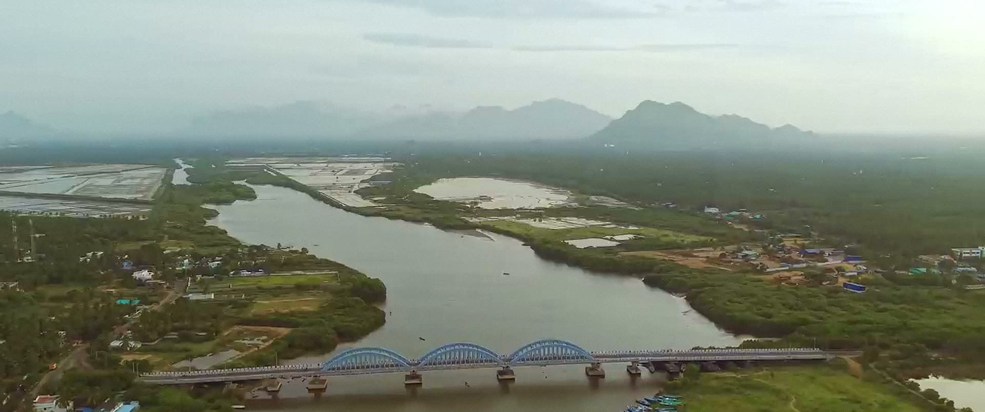 An aerial view of a wide river crossed by an arched bridge, with wetlands and hills beyond.
